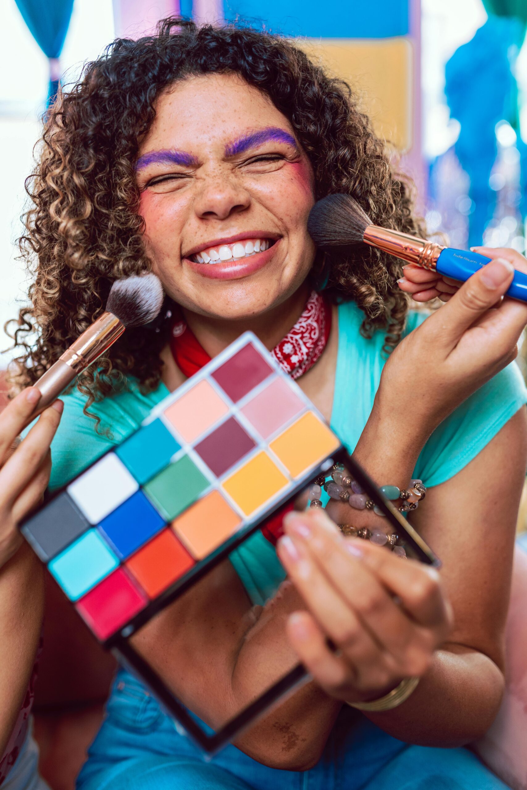 A cheerful woman with curly hair enjoys a colorful makeup session, smiling and holding a vibrant eyeshadow palette.